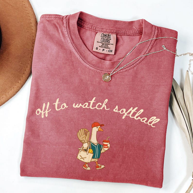May include: A dusty rose-colored t-shirt with the phrase "off to watch softball" in a cursive font. The shirt features a cartoon goose wearing a baseball uniform, holding a drink and a bag. A silver necklace with a pendant is also visible.