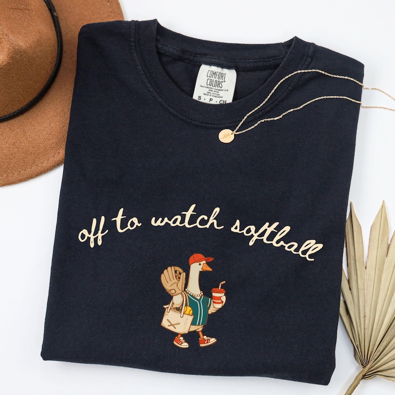 May include: Navy blue t-shirt with the phrase "off to watch softball" in cursive. The shirt features a cartoon goose wearing a baseball cap, holding a drink and a bag with a baseball glove. A gold necklace and a brown hat are also visible.