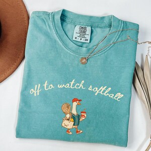 May include: Teal Comfort Colors t-shirt with the phrase "off to watch softball" in cursive. The shirt features a cartoon goose wearing a baseball cap, holding a drink and a bag with a baseball glove. A gold necklace is draped across the shirt.