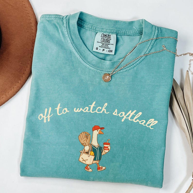 May include: Teal Comfort Colors t-shirt with the phrase "off to watch softball" in cursive. The shirt features a cartoon goose wearing a baseball cap, holding a drink and a bag with a baseball glove. A gold necklace is draped across the shirt.