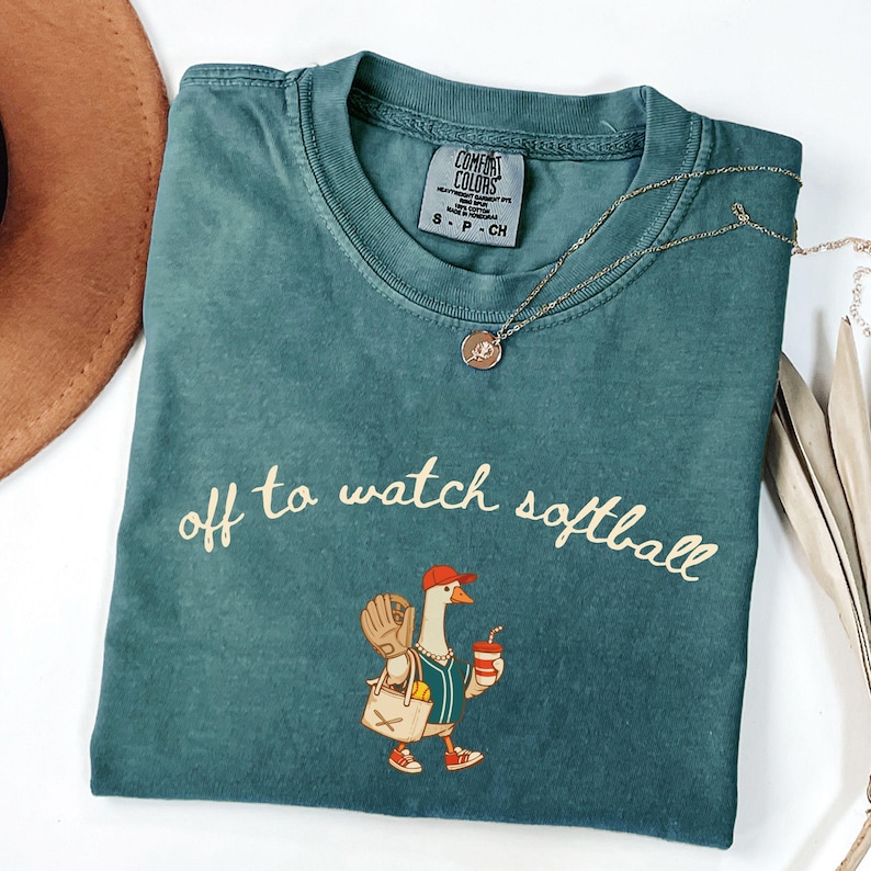 May include: Teal Comfort Colors t-shirt with the phrase "off to watch softball" in cursive. The shirt features a cartoon goose wearing a baseball cap, holding a drink and a baseball glove. A gold necklace is draped across the shirt.