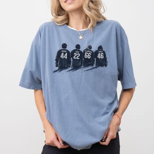 May include: A light blue t-shirt featuring a graphic of four figures with jersey numbers 44, 22, 66, and 46. The names Graham, Logan, Di Laurentis, and Tucker are above the numbers. The shirt has short sleeves and a crew neck.