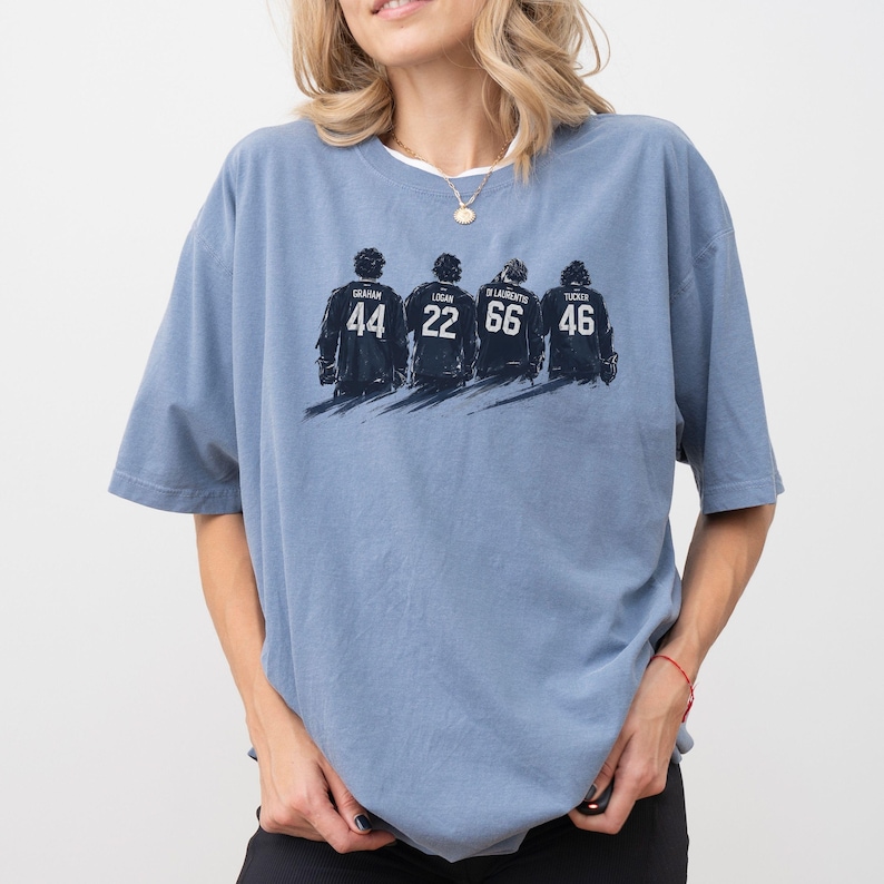 May include: A light blue t-shirt featuring a graphic of four figures with jersey numbers 44, 22, 66, and 46. The names Graham, Logan, Di Laurentis, and Tucker are above the numbers. The shirt has short sleeves and a crew neck.