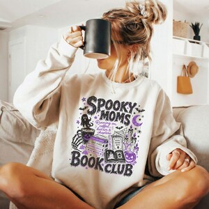 May include: A beige crewneck sweatshirt featuring a graphic print with the text "SPOOKY MOMS BOOK CLUB" and "Running on coffee, horror & chapters." The design includes illustrations of a ghost, books, candles, a pumpkin, and a spiderweb in black and purple.