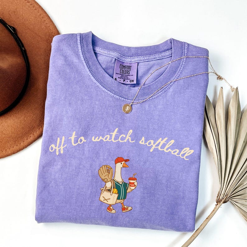 May include: A lilac Comfort Colors t-shirt with the phrase "off to watch softball" in cursive. The shirt features a cartoon goose wearing a baseball cap, holding a drink and a baseball glove. A gold necklace is draped across the shirt.