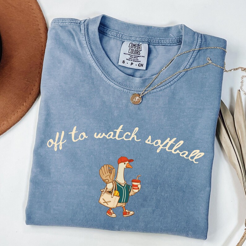 May include: A blue Comfort Colors t-shirt with the phrase "off to watch softball" in a cursive font. The shirt features a cartoon goose wearing a baseball cap, jersey, and carrying a bag and drink. A gold necklace is draped across the shirt.