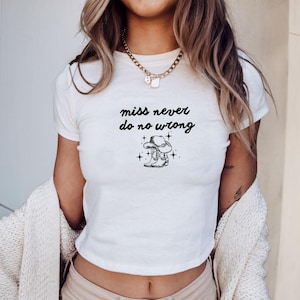 May include: White crop top with the text "miss never do no wrong" in black script. Below the text is a black and white graphic of a cowboy hat and boots with stars. The shirt is short-sleeved.