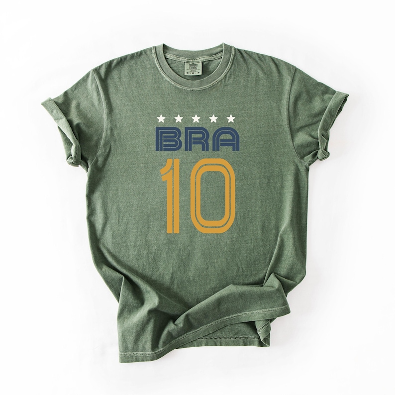 Peut inclure: T-shirt vert avec le mot "BRA" en bleu marine et le chiffre "10" en or. Au-dessus du mot "BRA" se trouvent cinq &eacute;toiles blanches. Le t-shirt a un col rond et des manches courtes.