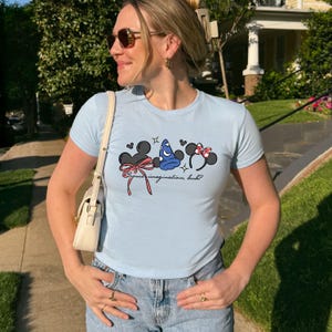 Puede incluir: Camiseta azul claro con tres dise&ntilde;os de orejas de rat&oacute;n de dibujos animados, lazos y un sombrero de mago. La camiseta tiene el texto "Some imagination, huh?" en una fuente cursiva. La camiseta se usa con vaqueros azules.