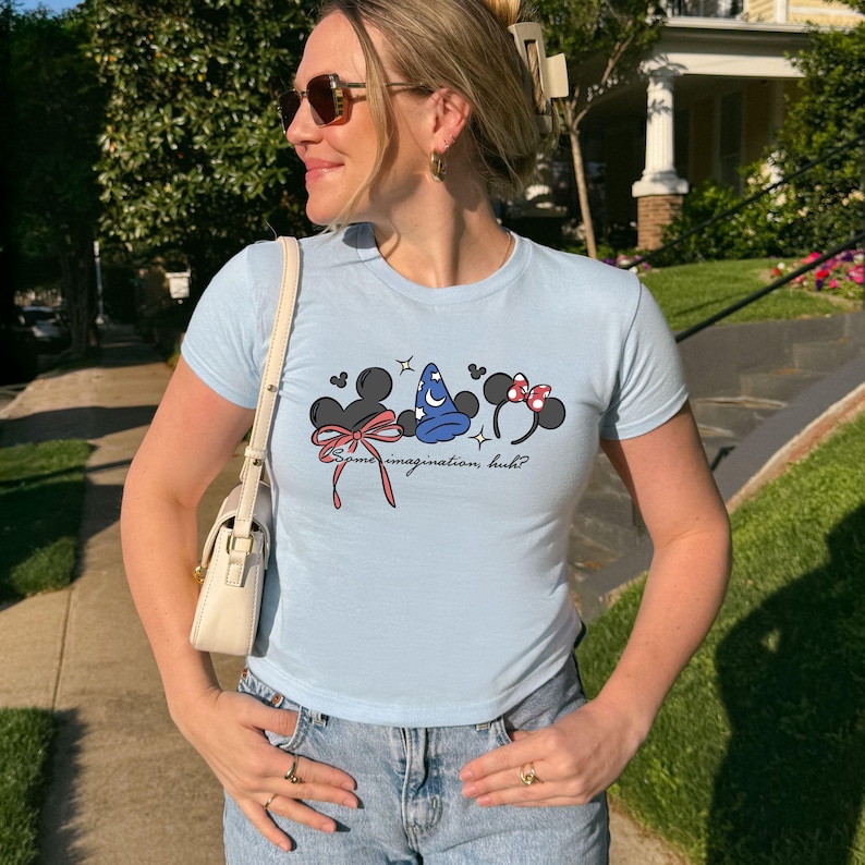 Puede incluir: Camiseta azul claro con tres dise&ntilde;os de orejas de rat&oacute;n de dibujos animados, lazos y un sombrero de mago. La camiseta tiene el texto "Some imagination, huh?" en una fuente cursiva. La camiseta se usa con vaqueros azules.