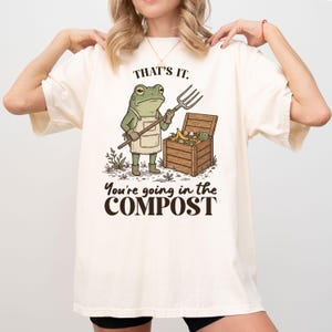 May include: Cream-colored t-shirt featuring a cartoon frog in an apron holding a pitchfork, standing next to a wooden compost bin. The text "THAT'S IT. You're going in the COMPOST" is printed above and below the frog illustration.