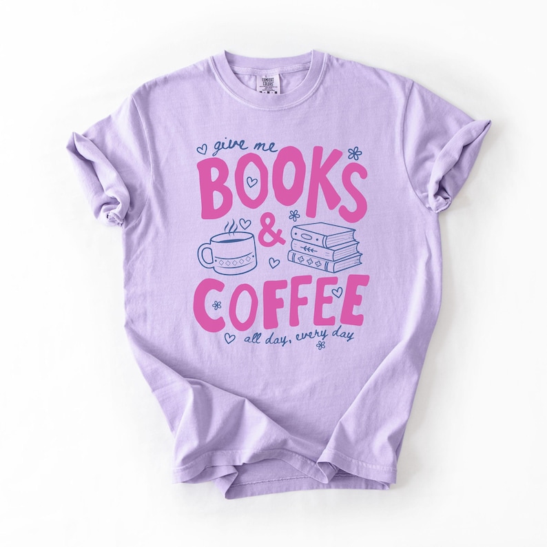 May include: A light purple short-sleeve t-shirt featuring the pink text "BOOKS & COFFEE" with blue line art illustrations of a steaming mug and a stack of books. The design includes small blue hearts and flowers, with the text "give me" and "all day, every day".