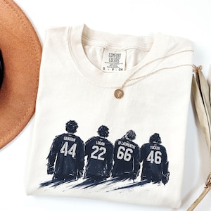 May include: Cream-colored Comfort Colors t-shirt featuring a graphic of four figures in dark blue jerseys with names and numbers: Graham 44, Logan 22, Di Laurentis 66, and Tucker 46. A gold necklace and a brown hat are also visible.