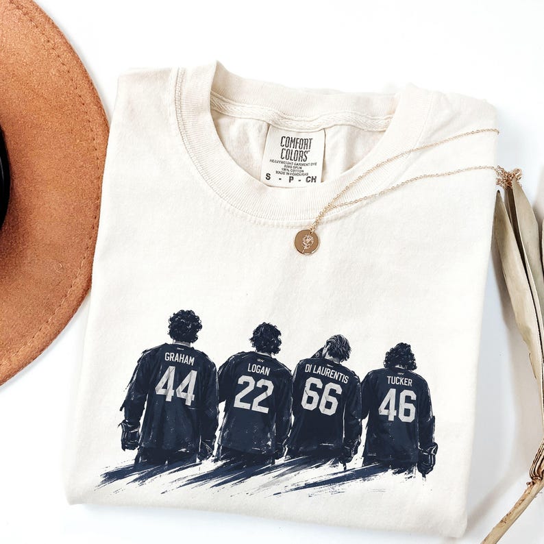 May include: Cream-colored Comfort Colors t-shirt featuring a graphic of four figures in dark blue jerseys with names and numbers: Graham 44, Logan 22, Di Laurentis 66, and Tucker 46. A gold necklace and a brown hat are also visible.