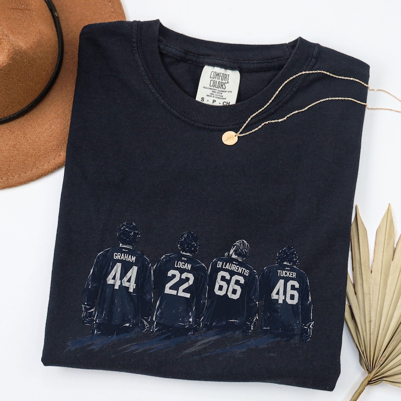 May include: Navy blue t-shirt featuring a graphic of four hockey players with their jersey numbers: 44, 22, 66, and 46. The shirt includes the "Comfort Colors" tag. A gold necklace and a brown hat are also visible.