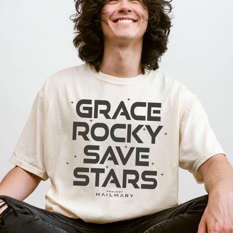 May include: Cream-colored t-shirt with the words "GRACE ROCKY SAVE STARS" in bold, black letters. The text is arranged vertically and is accompanied by small cross symbols. The shirt also features the text "PROJECT HAILMARY" at the bottom.