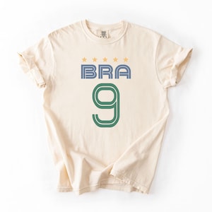 Peut inclure: T-shirt cr&egrave;me avec le mot "BRA" en bleu et le chiffre "9" en vert. Au-dessus du mot "BRA" se trouvent quatre &eacute;toiles dor&eacute;es. Le t-shirt a des manches courtes et un col rond.