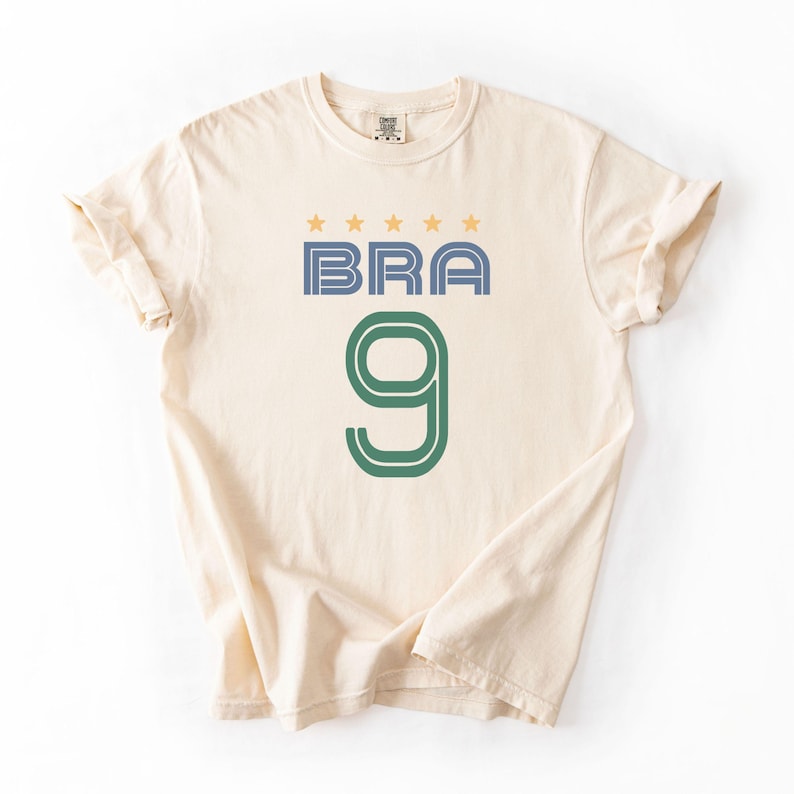 Peut inclure: T-shirt cr&egrave;me avec le mot "BRA" en bleu et le chiffre "9" en vert. Au-dessus du mot "BRA" se trouvent quatre &eacute;toiles dor&eacute;es. Le t-shirt a des manches courtes et un col rond.