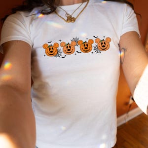May include: A white short-sleeve t-shirt featuring a horizontal graphic print of four orange pumpkins with mouse-ear shapes, black facial features, and green stems, interspersed with black spider web designs, small stars, and flying bat silhouettes.
