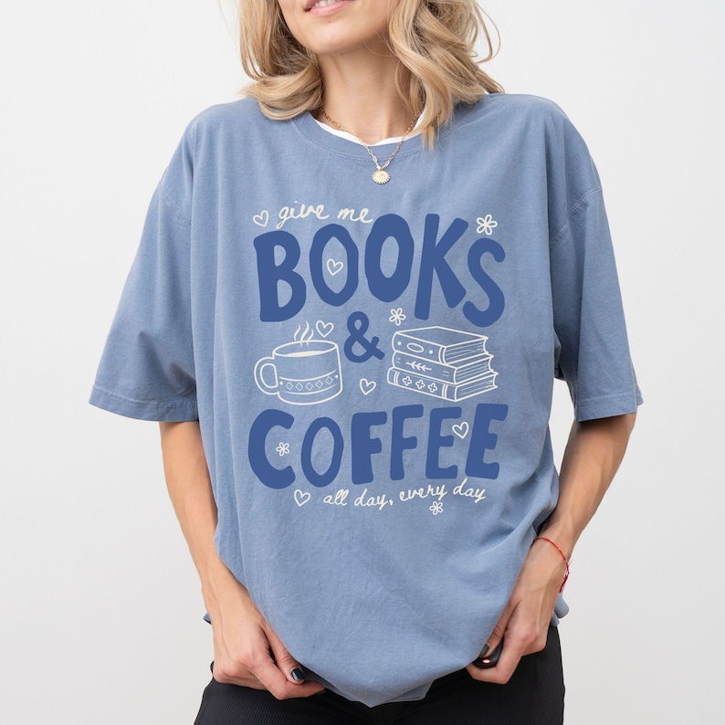 May include: A dusty blue short-sleeve t-shirt featuring dark blue text that reads "give me BOOKS & COFFEE all day, every day". The graphic includes a white line drawing of a coffee mug and a stack of three books, accented with small white hearts and flowers.