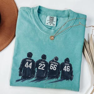 May include: A teal Comfort Colors t-shirt featuring a graphic of four hockey players with their jersey numbers: 44, 22, 66, and 46. A gold necklace with a pendant rests on the shirt. The shirt is folded, with a brown hat visible in the background.