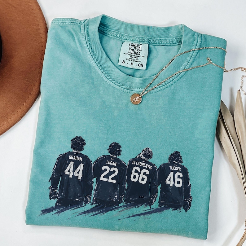 May include: A teal Comfort Colors t-shirt featuring a graphic of four hockey players with their jersey numbers: 44, 22, 66, and 46. A gold necklace with a pendant rests on the shirt. The shirt is folded, with a brown hat visible in the background.