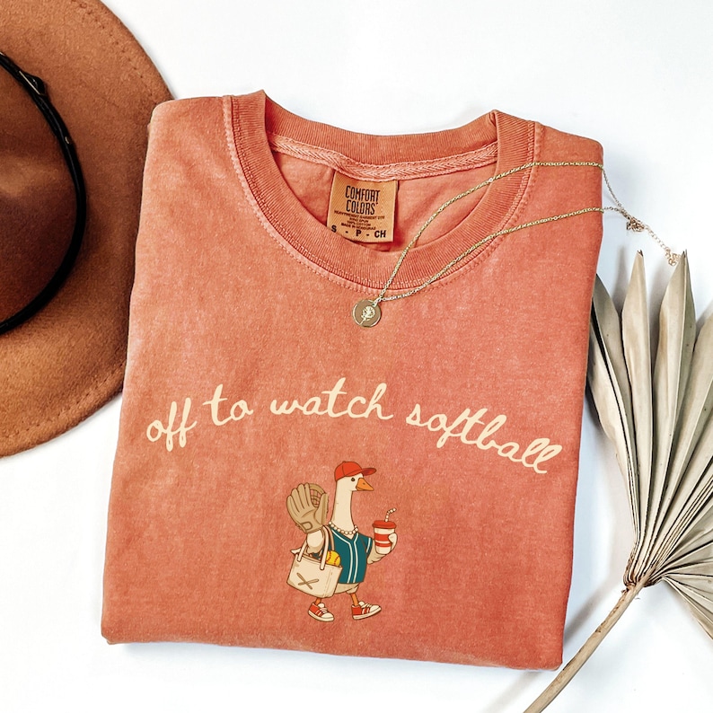 May include: Coral-colored Comfort Colors t-shirt with the phrase "off to watch softball" in white script. The shirt features a cartoon goose wearing a baseball cap, holding a coffee cup and a baseball glove. A gold necklace is draped across the shirt.