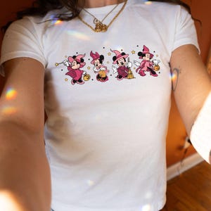 May include: A white short-sleeved t-shirt featuring a graphic print of four Minnie Mouse characters dressed in pink witch costumes. The characters are depicted with wands, broomsticks, and pumpkin buckets, surrounded by small yellow stars.