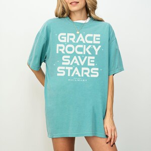 May include: A turquoise t-shirt with the words "GRACE ROCKY SAVE STARS" in large white block letters. The text "PROJECT HAILMARY" is printed below. The shirt is a relaxed fit and appears to be made of soft material.