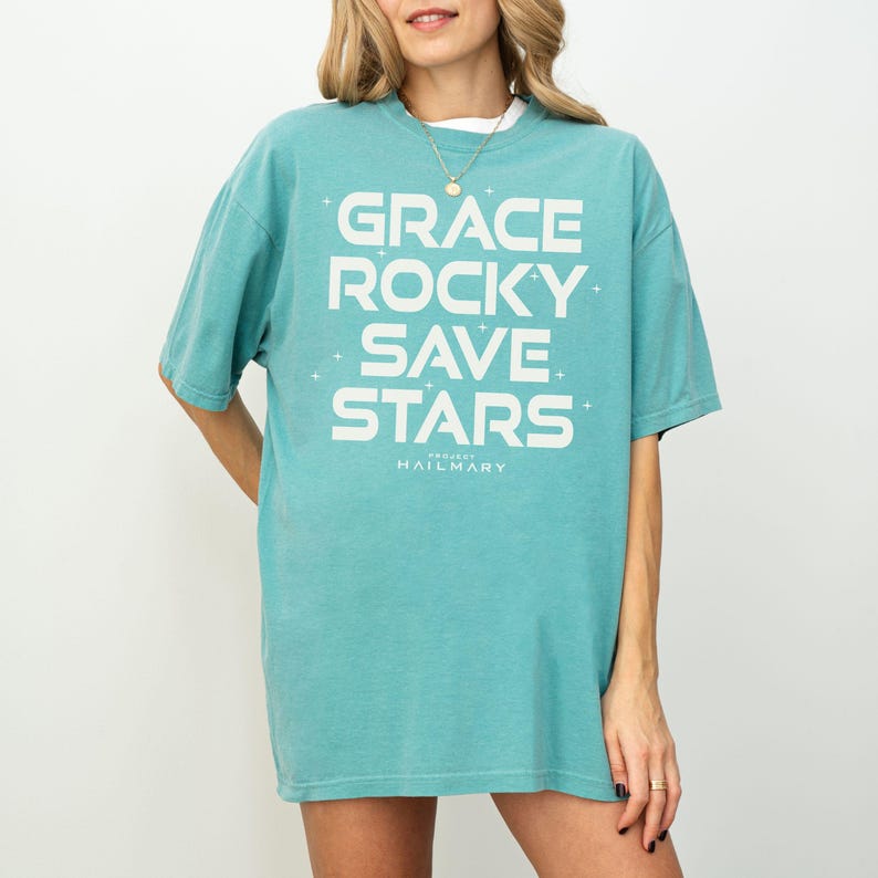 May include: A turquoise t-shirt with the words "GRACE ROCKY SAVE STARS" in large white block letters. The text "PROJECT HAILMARY" is printed below. The shirt is a relaxed fit and appears to be made of soft material.