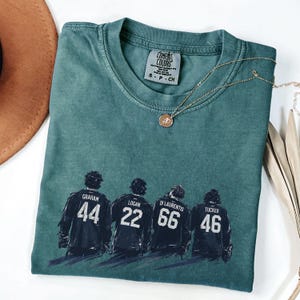May include: Teal Comfort Colors t-shirt with a graphic of four hockey players' backs, each with a name and number: Graham 44, Logan 22, Di Laurentis 66, and Tucker 46. A gold necklace with a pendant rests on the shirt.