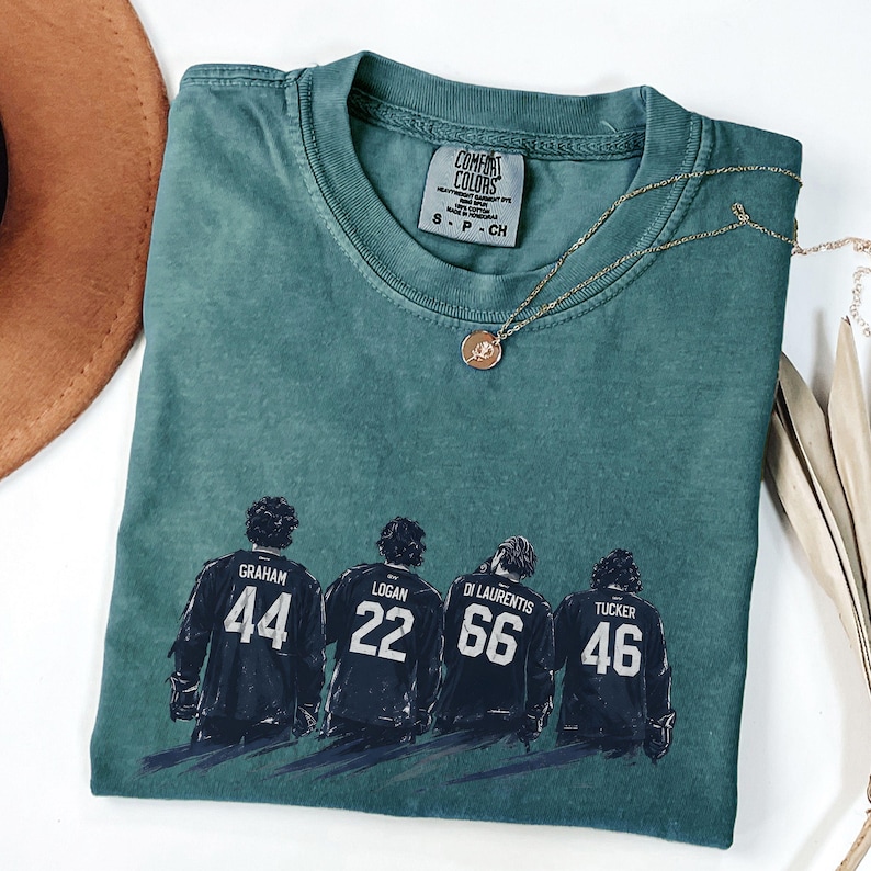 May include: Teal Comfort Colors t-shirt with a graphic of four hockey players' backs, each with a name and number: Graham 44, Logan 22, Di Laurentis 66, and Tucker 46. A gold necklace with a pendant rests on the shirt.