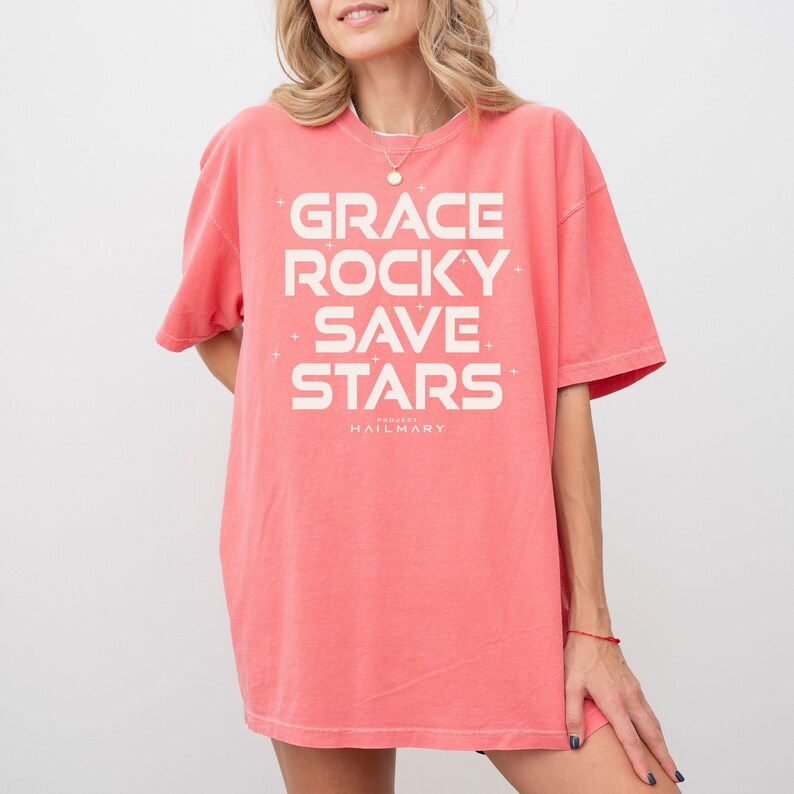 May include: Coral-colored t-shirt with the words "GRACE ROCKY SAVE STARS" in white block letters. The text is arranged vertically, with the word "HAILMARY" below. The shirt has short sleeves and a relaxed fit.