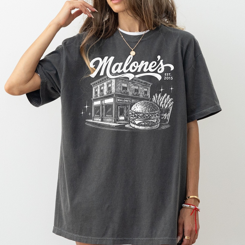 K&ouml;nnte beinhalten: Dunkelgraues T-Shirt mit einer Grafik eines Restaurants namens "Malone's", einem Burger und Pommes Frites. Das Design enth&auml;lt den Text "Malone's" in einer Schreibschrift, den Namen des Unternehmens und das Jahr "2015".