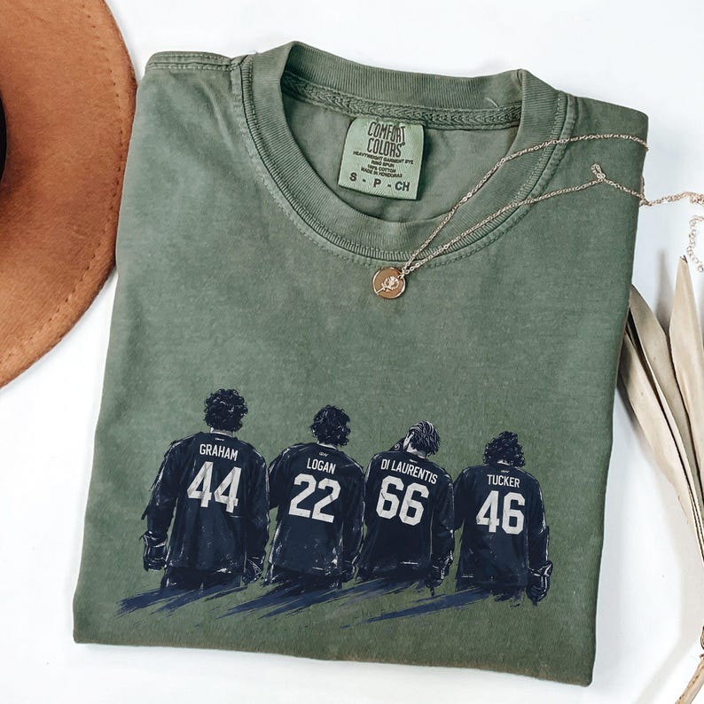 May include: Olive green Comfort Colors t-shirt featuring four figures in hockey jerseys with the names Graham, Logan, Di Laurentis, and Tucker, and the numbers 44, 22, 66, and 46. A gold necklace with a pendant is on the shirt.
