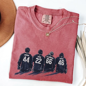 May include: A red Comfort Colors t-shirt with a graphic of four hockey players with their jersey numbers: 44, 22, 66, and 46. The shirt has a crew neck and a small tag. A gold necklace with a pendant is draped across the shirt.