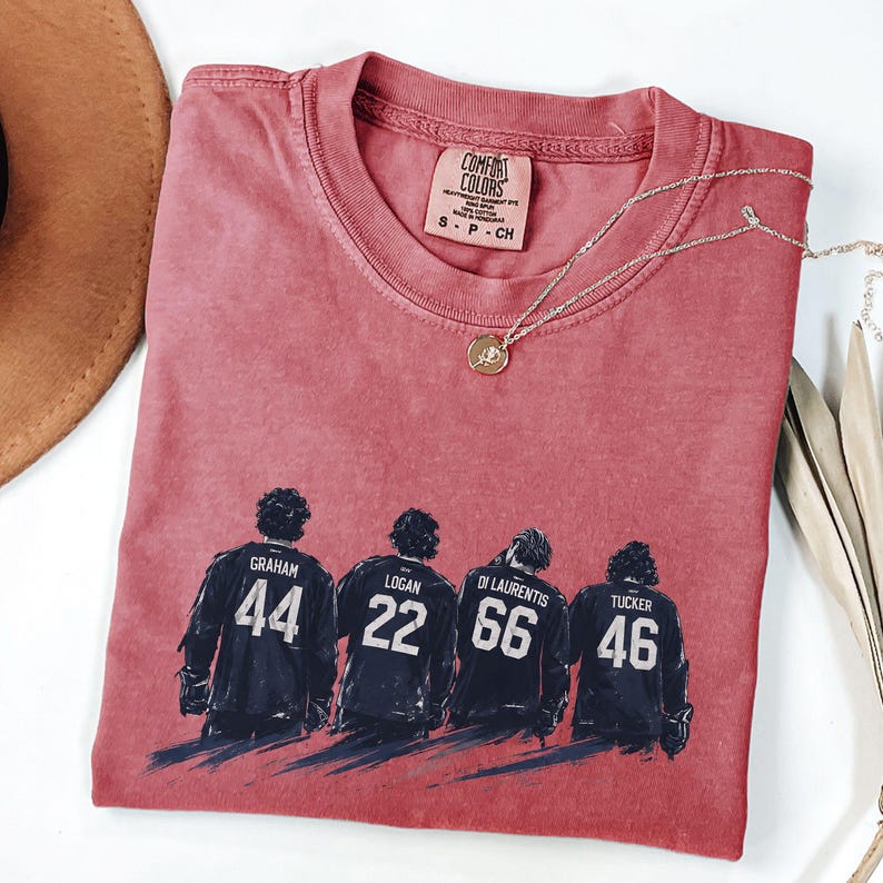 May include: A red Comfort Colors t-shirt with a graphic of four hockey players with their jersey numbers: 44, 22, 66, and 46. The shirt has a crew neck and a small tag. A gold necklace with a pendant is draped across the shirt.
