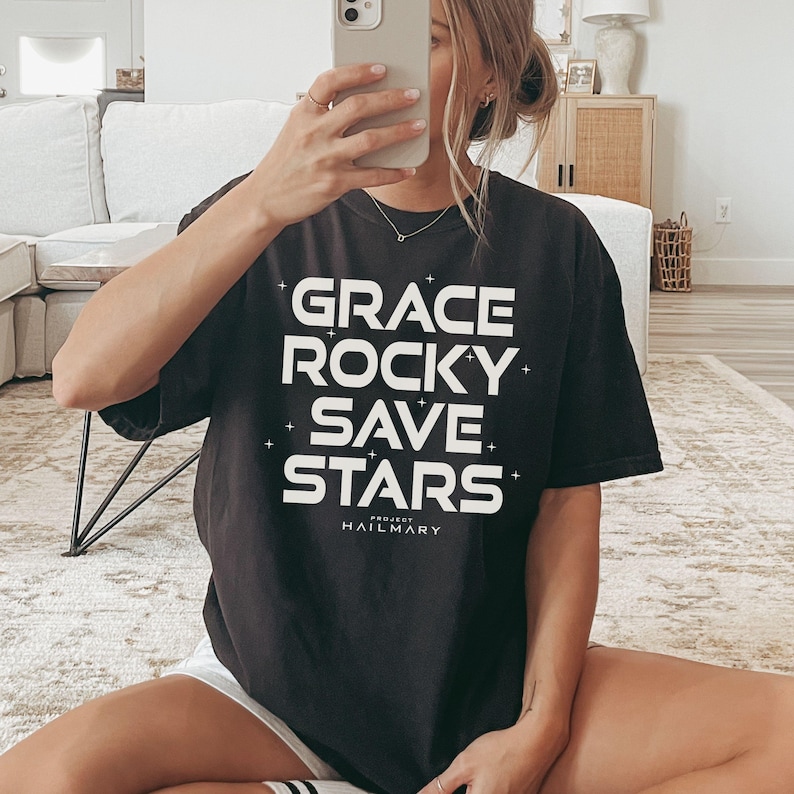 May include: Black t-shirt with the words "GRACE ROCKY SAVE STARS" in large white block letters. The text is arranged vertically with the word "PROJECT HAILMARY" printed below. The shirt is a dark color and appears to be made of soft material.