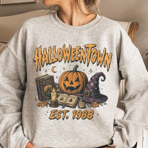 May include: A light gray crewneck sweatshirt featuring a graphic print with the text HALLOWEENTOWN EST. 1988. The design includes a jack-o'-lantern, a purple witch hat, an open book, a closed book, a lit candle, and a small potion bottle.