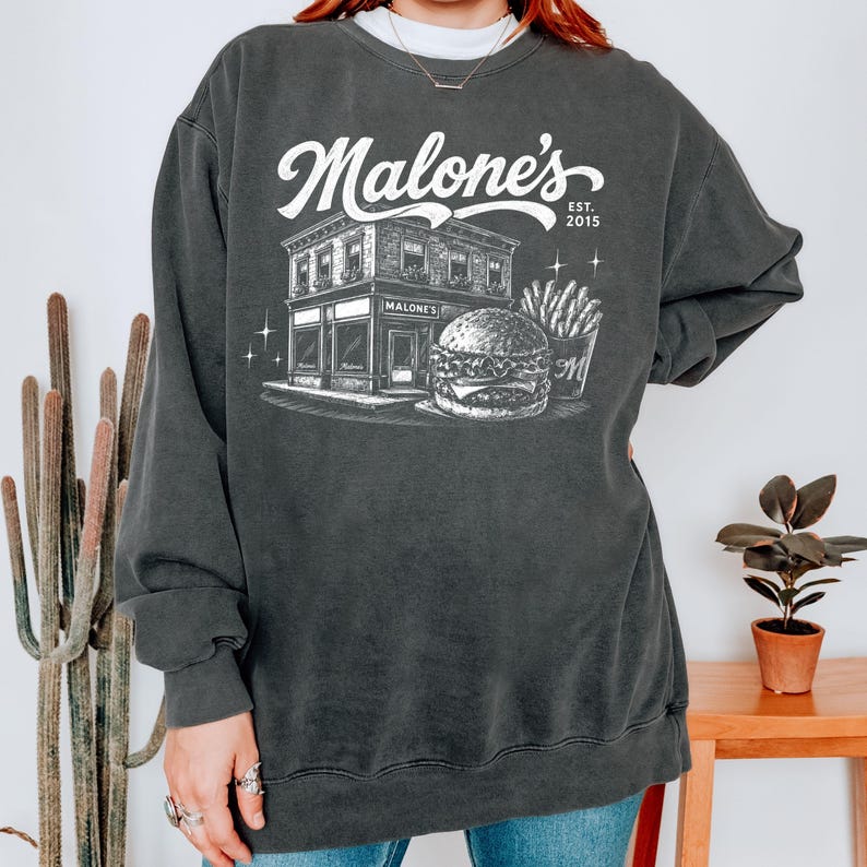 K&ouml;nnte beinhalten: Dunkelgraues Sweatshirt mit einem wei&szlig;en Aufdruck eines Restaurants namens "Malone's" und dem Text "Est. 2015". Die Grafik zeigt einen Burger, Pommes Frites und das Restaurantgeb&auml;ude.