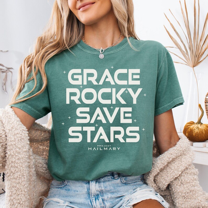 May include: A teal-colored t-shirt with the words "GRACE ROCKY SAVE STARS" in large white block letters. The text is slightly distressed, and the shirt has a relaxed fit. The words "PROJECT HAILMARY" are printed below the main text.