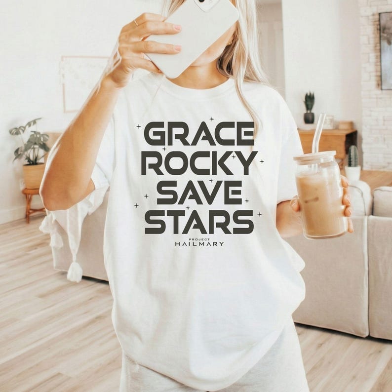 May include: White t-shirt with the words "GRACE ROCKY SAVE STARS" in a bold, dark gray font. The text is stacked vertically. Below the text is the text "PROJECT HAILMARY" in a smaller font. The shirt is a casual, everyday wear item.