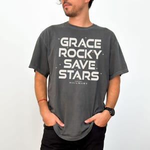 May include: A dark gray t-shirt with the words "GRACE ROCKY SAVE STARS" in large white block letters. The text is arranged vertically with the word "HAILMARY" below. The shirt is a short-sleeved, casual style.