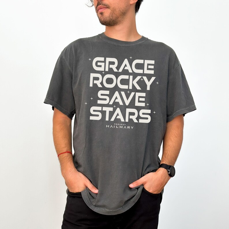May include: A dark gray t-shirt with the words "GRACE ROCKY SAVE STARS" in large white block letters. The text is arranged vertically with the word "HAILMARY" below. The shirt is a short-sleeved, casual style.