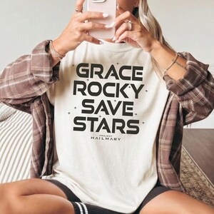 May include: Cream-colored t-shirt with the words "GRACE ROCKY SAVE STARS" in bold black letters. The text is centered, with small cross symbols on either side of the words. The brand name "PROJECT HAILMARY" is printed below the text.