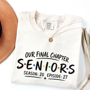 May include: A cream-colored t-shirt with black text that reads "OUR FINAL CHAPTER SENIORS SEASON: 20 EPISODE: 27" with colorful dots between the letters of the word "SENIORS". The shirt features a Comfort Colors brand tag at the neck.