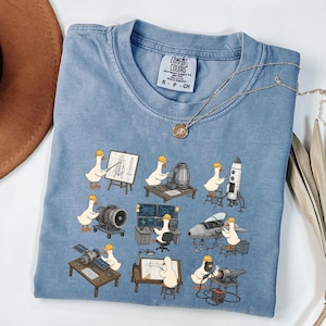 May include: A light blue Comfort Colors t-shirt featuring a whimsical design of cartoon geese in construction hats working on various engineering projects, including rockets and blueprints. A gold necklace with a pendant is also visible.