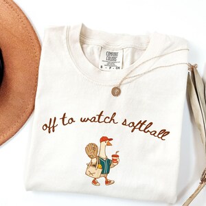 May include: Cream-colored t-shirt with the phrase "off to watch softball" in brown script. The shirt features a cartoon goose wearing a baseball cap, holding a drink and a baseball glove. A gold necklace and a brown hat are also visible.