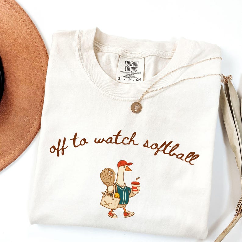 May include: Cream-colored t-shirt with the phrase "off to watch softball" in brown script. The shirt features a cartoon goose wearing a baseball cap, holding a drink and a baseball glove. A gold necklace and a brown hat are also visible.