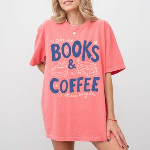 May include: A coral-colored short-sleeve t-shirt featuring the blue text "give me BOOKS & COFFEE" and "all day, every day". The design includes white line art of a coffee mug and a stack of three books, accented with small hearts and flowers.